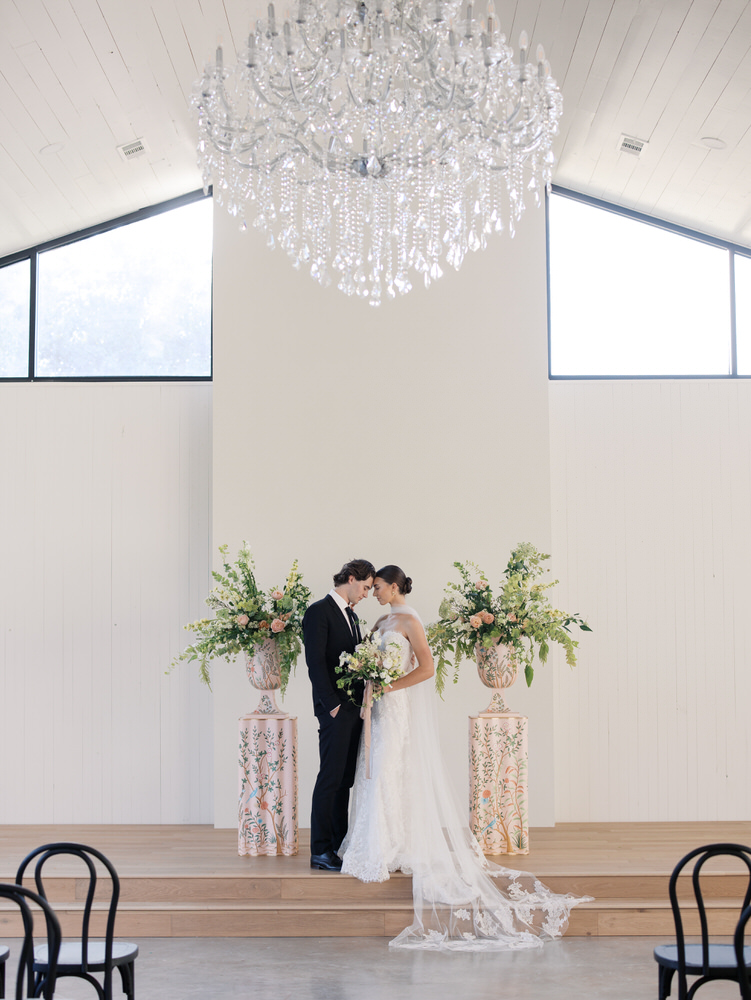 Bride in white lace gown and groom in black suit stand forehead to forehead holding a bouquet, flanked by two tall floral arrangements inside a bright venue with a large crystal chandelier overhead.