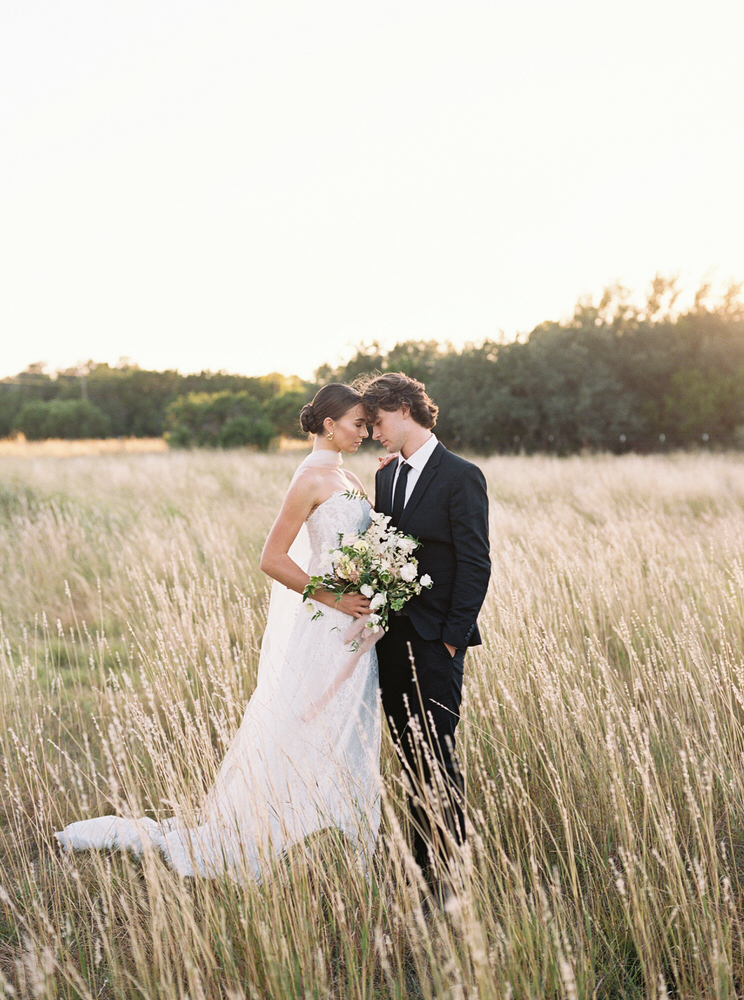 Bride in white dress holding a bouquet standing closely forehead-to-forehead with groom in black suit in a sunlit grassy field.