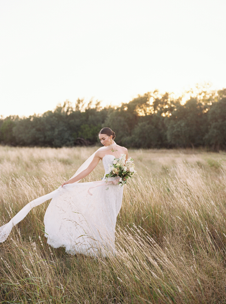 Bride in a white strapless wedding gown holding a bouquet, standing in a sunlit grassy field at sunset.
