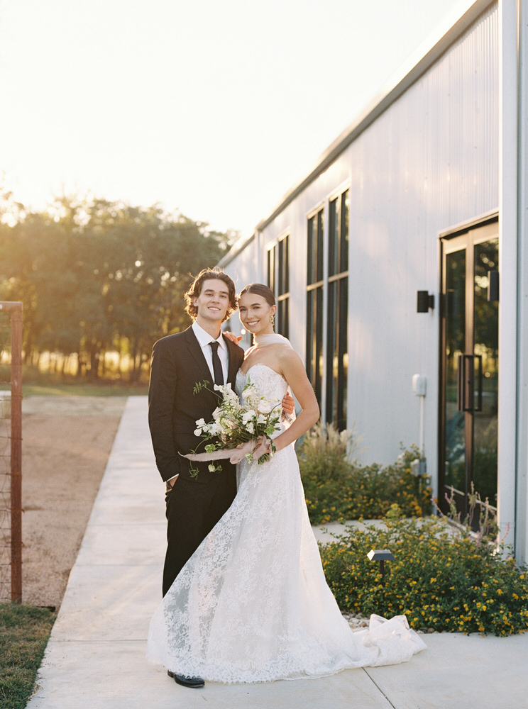 Smiling bride in lace wedding dress holding a bouquet and groom in black suit posing outside near a modern building at sunset.