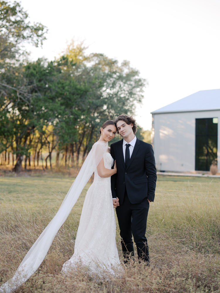 Bride in a white lace gown with a long veil and groom in a black suit and tie standing hand in hand in a grassy field with trees and a white building in the background.