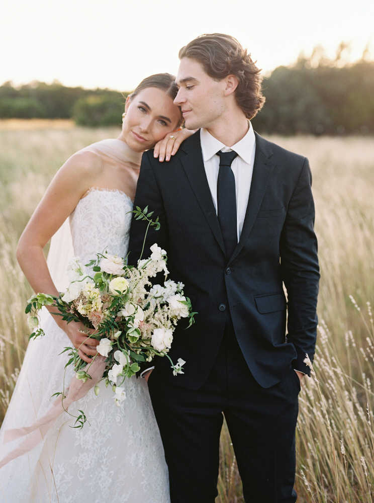 Bride in white lace dress holding a bouquet leans her head on the groom's shoulder in a field at sunset.