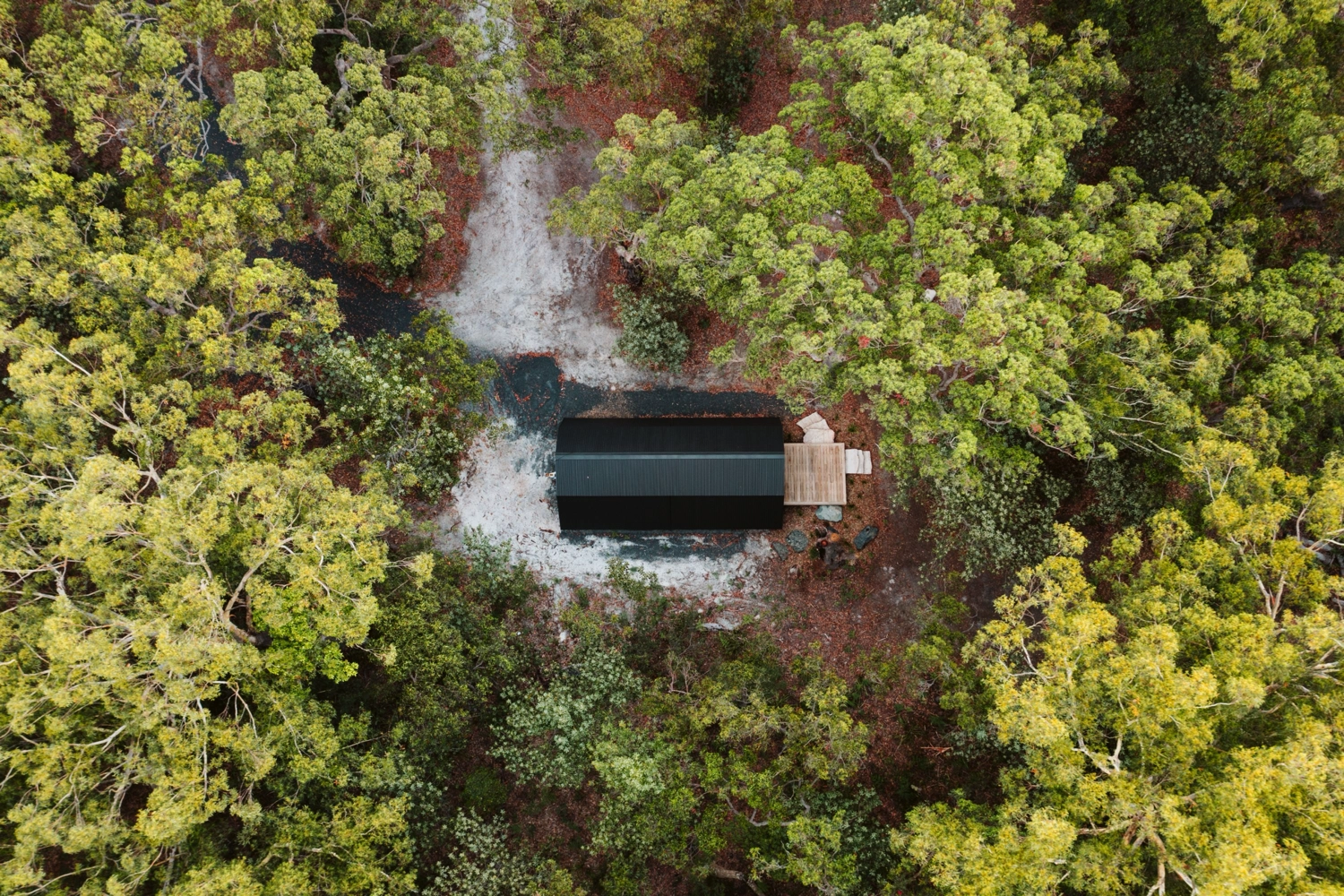 Aerial view of The Hideaway, a tiny studio with a black roof, nestled among dense green trees and a dirt clearing, featuring a wooden deck and scattered outdoor furniture—perfect for a romantic getaway.