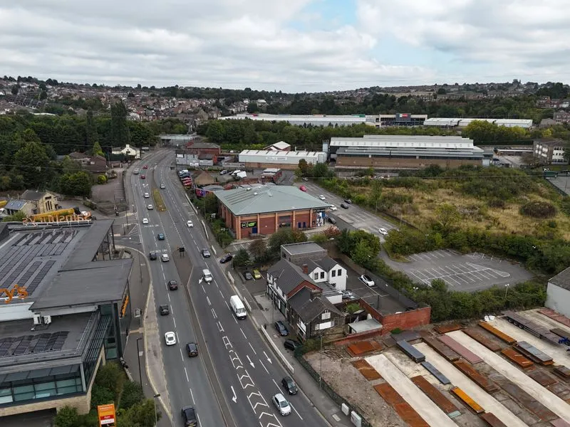 Aerial view of a road with cars, adjacent buildings, parking lots, greenery, and residential houses in the background under a cloudy sky.