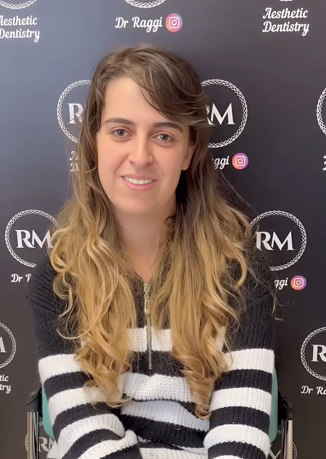 Smiling woman with long wavy blonde hair wearing a black and white striped sweater sitting in front of a black backdrop with 'Dr Raggi Aesthetic Dentistry' logos.