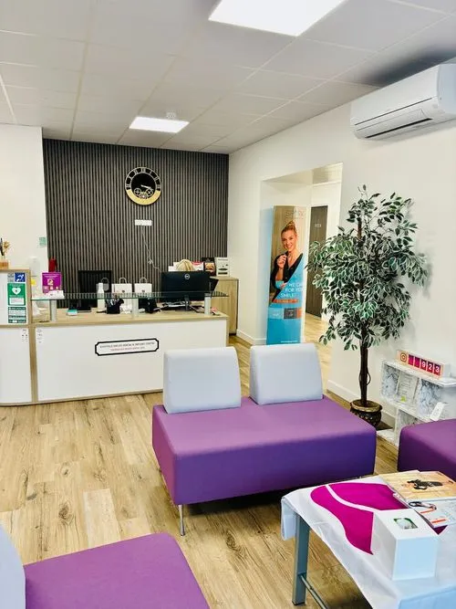 Modern waiting room with purple cushioned chairs, wooden floor, a reception desk, wall clock, and a potted plant.