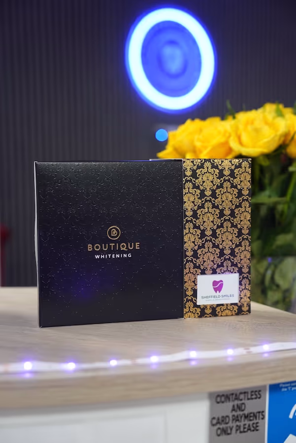 Black and gold decorative Boutique Whitening box on a counter with yellow flowers and a neon blue circular light in the background.