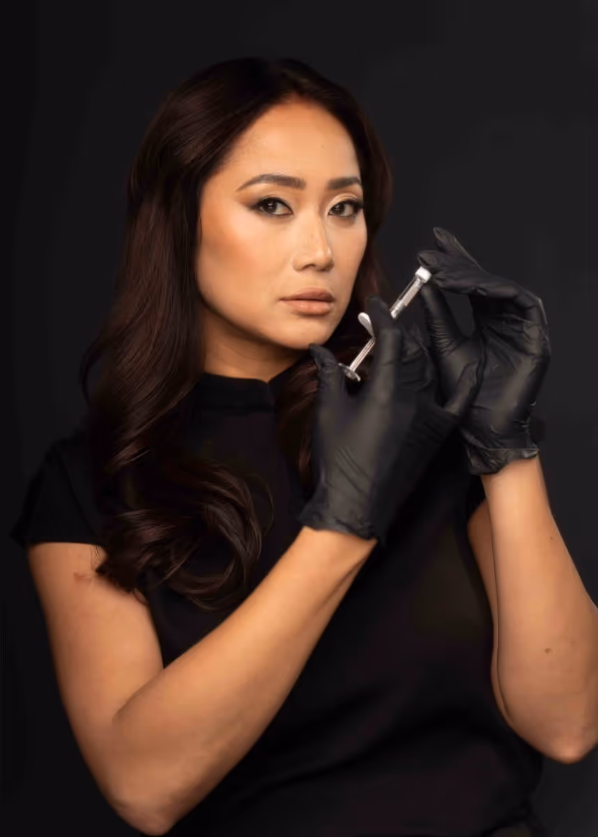 Woman with long dark hair wearing black gloves, holding a syringe against a black background.