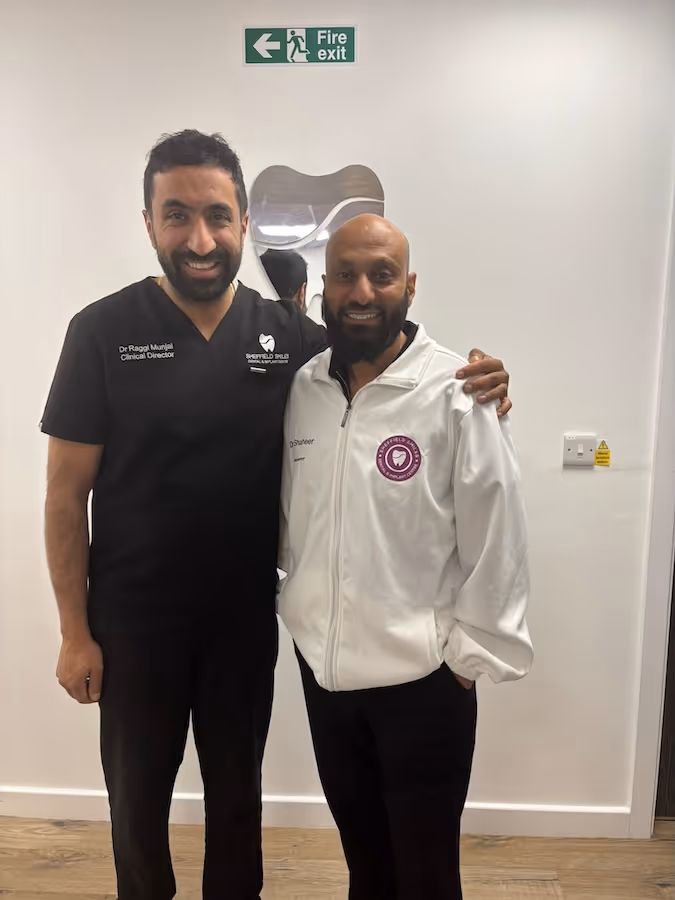 Two smiling men standing side by side indoors, one wearing a black clinical uniform and the other a white jacket, with a fire exit sign on the wall above them.