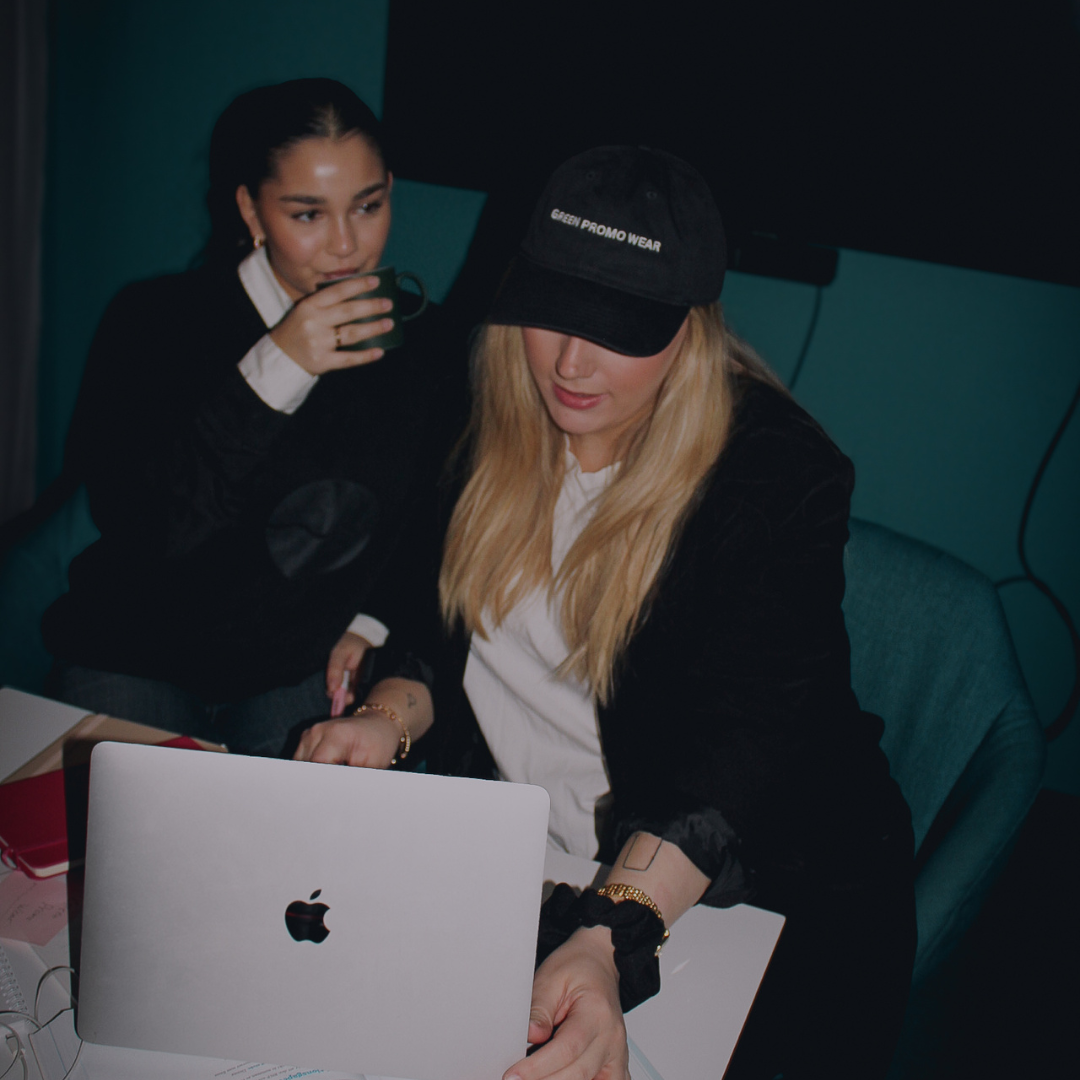 Two women working together at a table with a laptop, one drinking from a cup and the other wearing a black cap reading 'GREEN PROMO WEAR'.