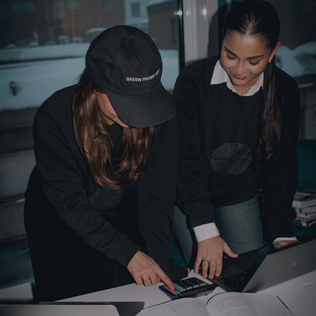 Two women working together at a desk with a calculator, laptop, and open book, one pointing at the calculator.