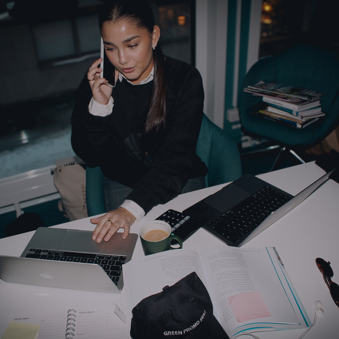 Woman sitting at a desk with two laptops, talking on a smartphone, with an open book, a coffee cup, and a black cap labeled 'GREEN PROMO WEAR'.