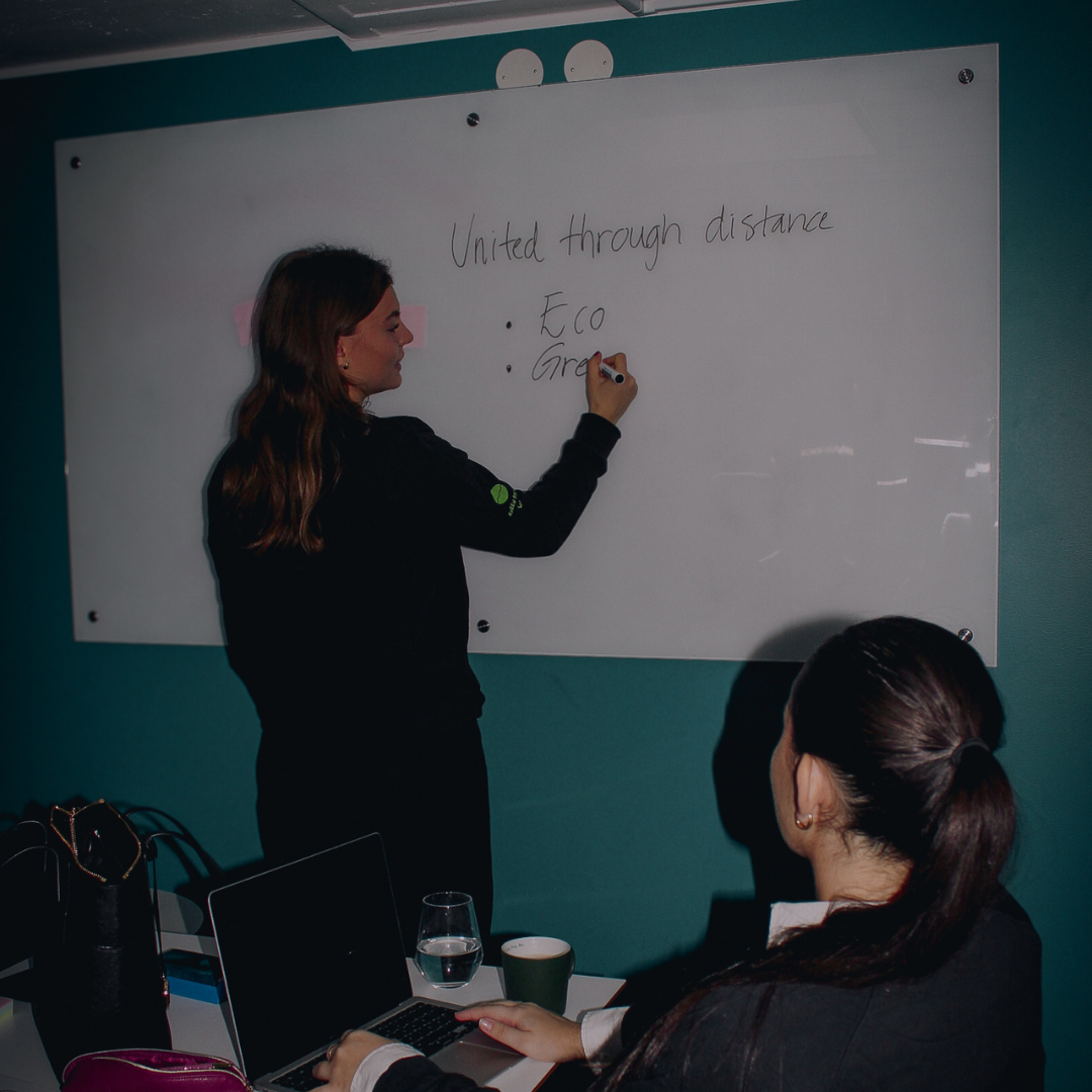 Woman writing on a whiteboard with the text 'United through distance' while another woman watches seated with a laptop and a glass of water on the table.