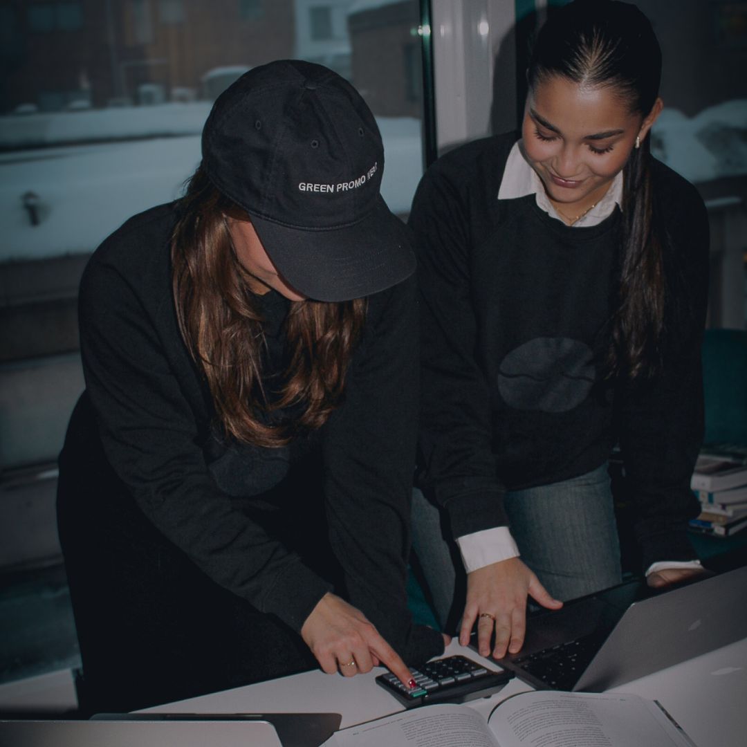 Two women working together at a desk with a calculator, laptop, and open book, one pointing at the calculator.