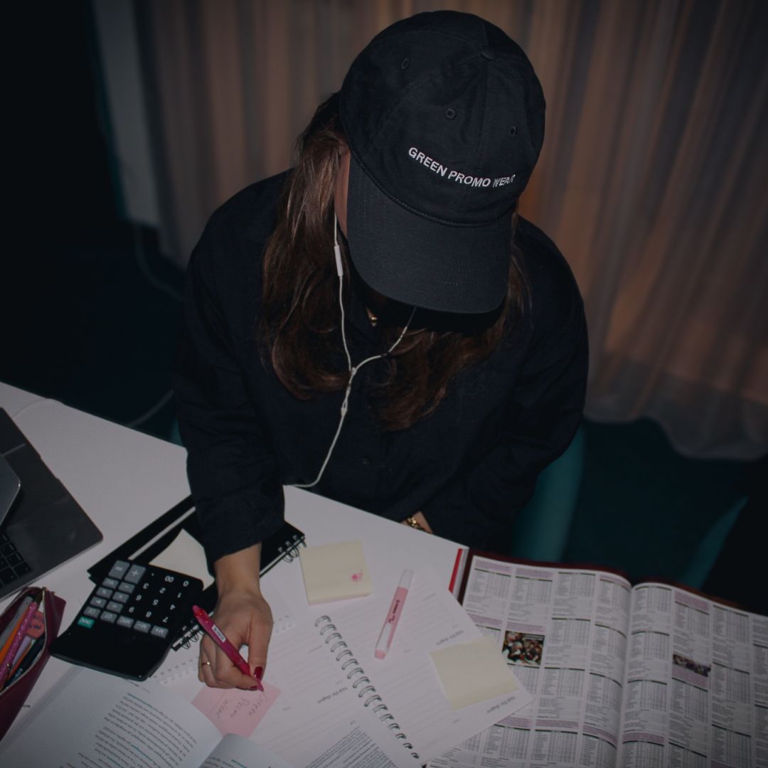 A person wearing a black cap and dark shirt is writing with a pink pen on a sticky note at a cluttered desk with notebooks, papers, a calculator, and a laptop.