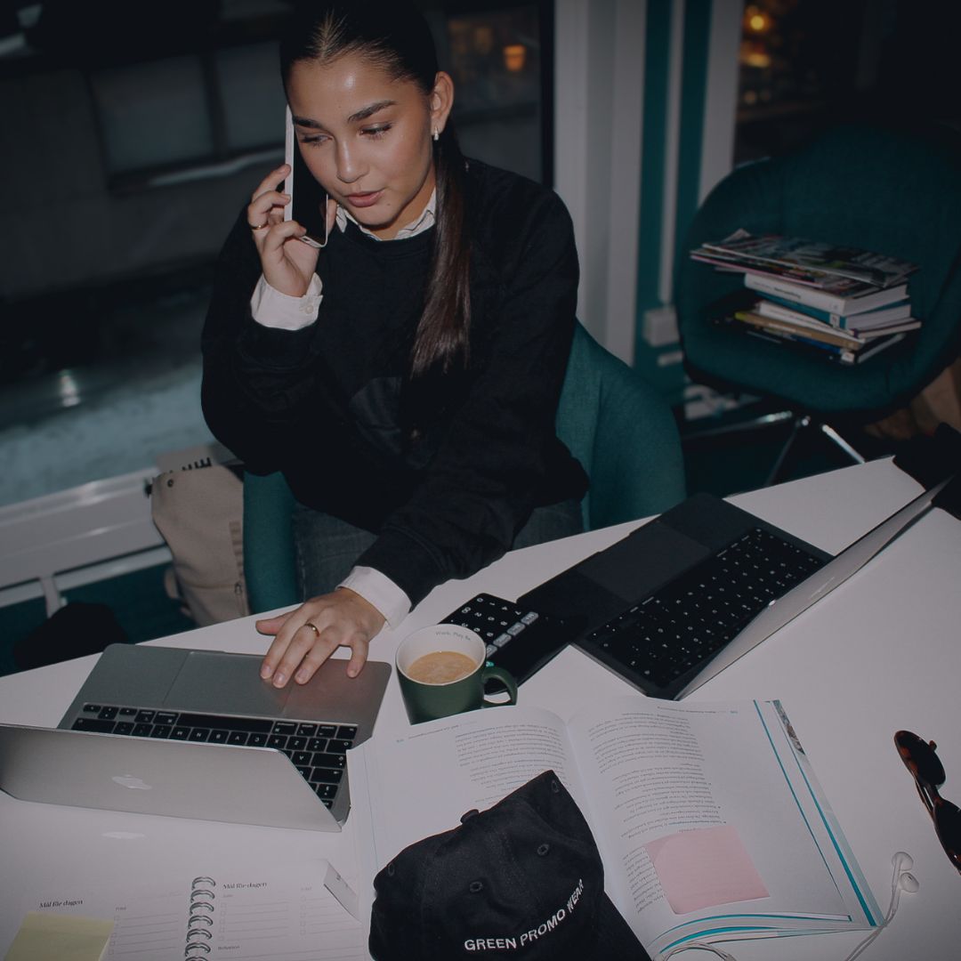 Woman sitting at a desk with two laptops, talking on a smartphone, with an open book, a coffee cup, and a black cap labeled 'GREEN PROMO WEAR'.
