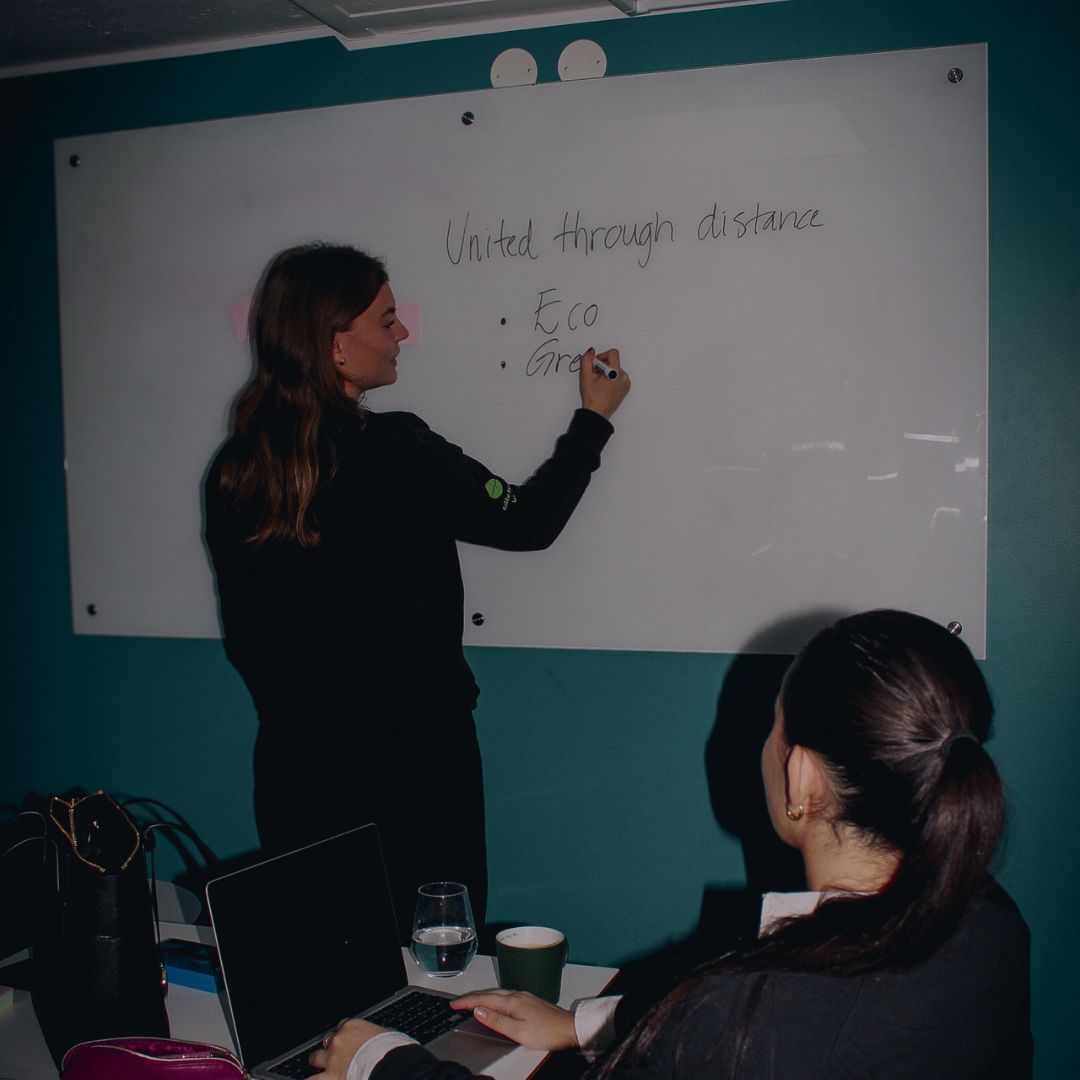 Woman writing on a whiteboard with the text 'United through distance' while another woman watches seated with a laptop and a glass of water on the table.