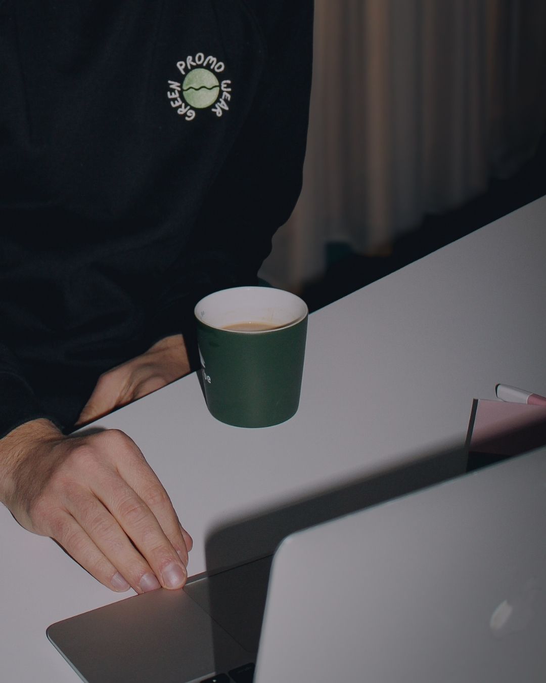 Person wearing a black sweatshirt with a green logo working on a laptop next to a green coffee cup on a white table.
