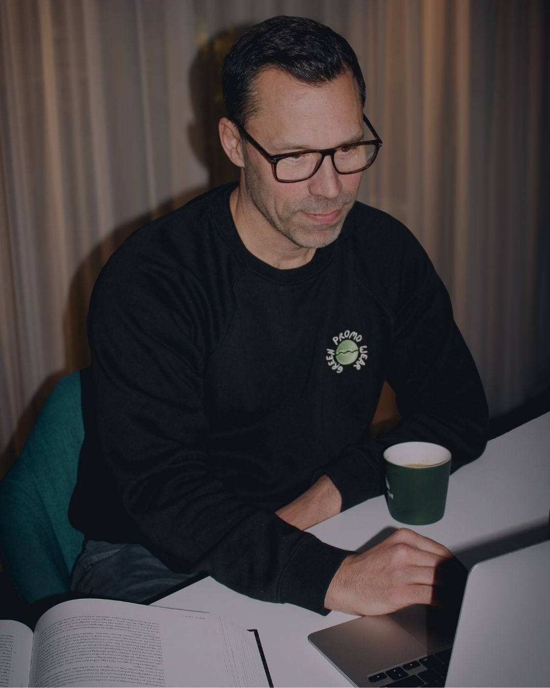Man wearing glasses and a black sweatshirt working on a laptop at a white desk with an open book and a green coffee cup.