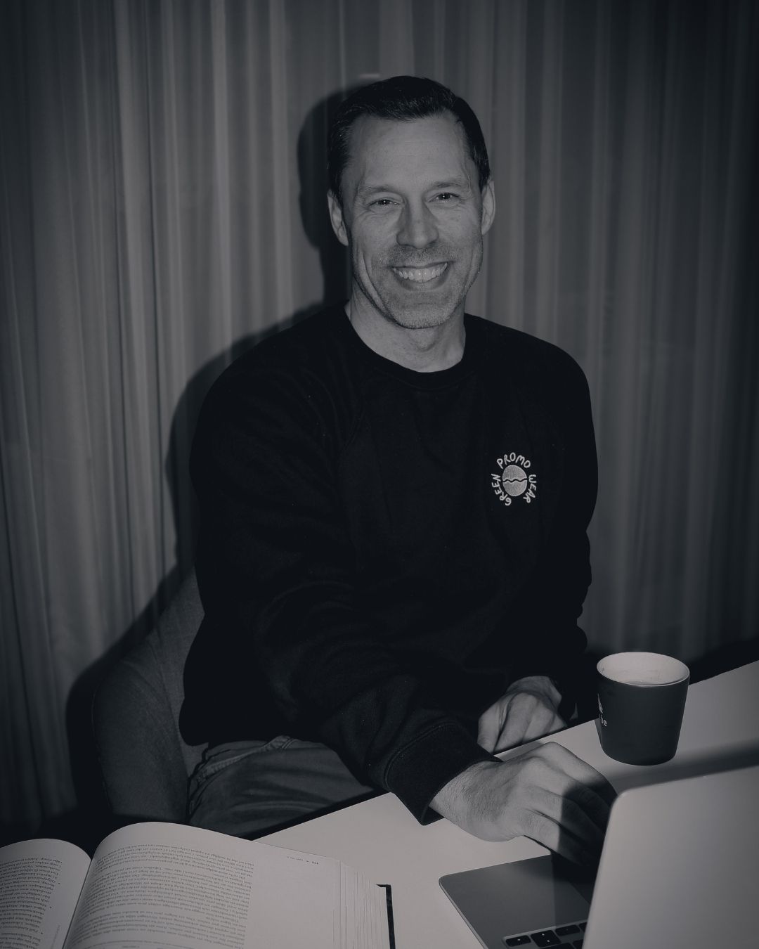 Smiling man sitting at a desk with an open book, using a laptop and a coffee cup nearby.