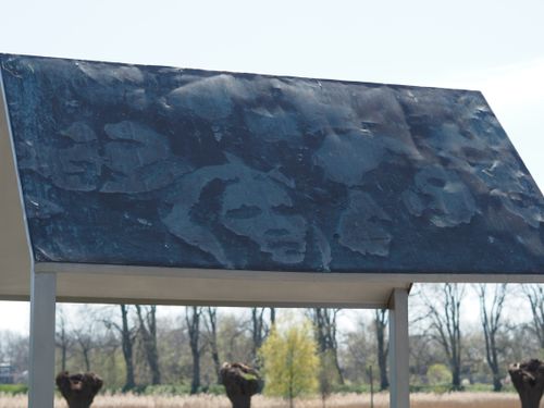 Foto van de schuilplaats, onderdeel van het monument. Het zijn vier palen met een puntdak er op. Op het dak zie je gezichten