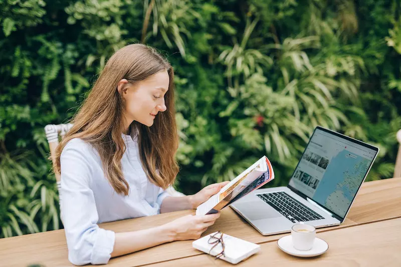 A lady is reading book in front on the laptop 