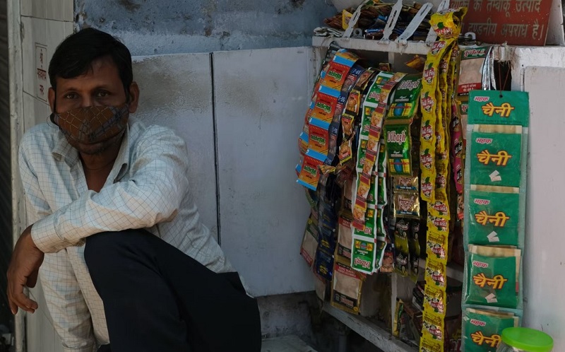 A Man Wearing Face Mask Sitting At Pan Parlour and Some Products Are Hanging on Tables