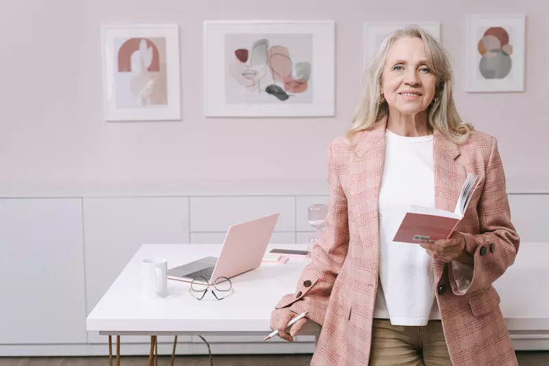 An elderly woman in pink blazer smiling while holding a book 