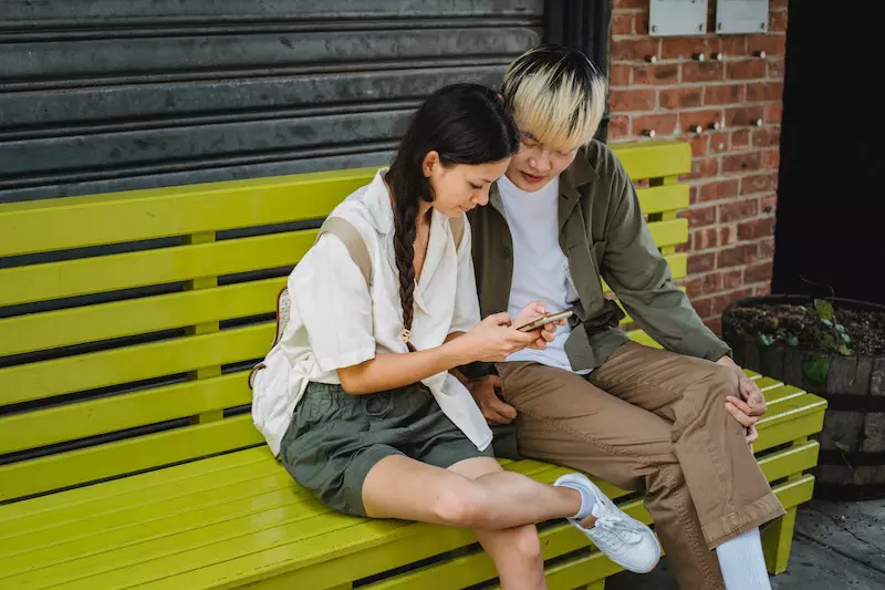A girl and boy is using smart phone sitting on the yellow bench