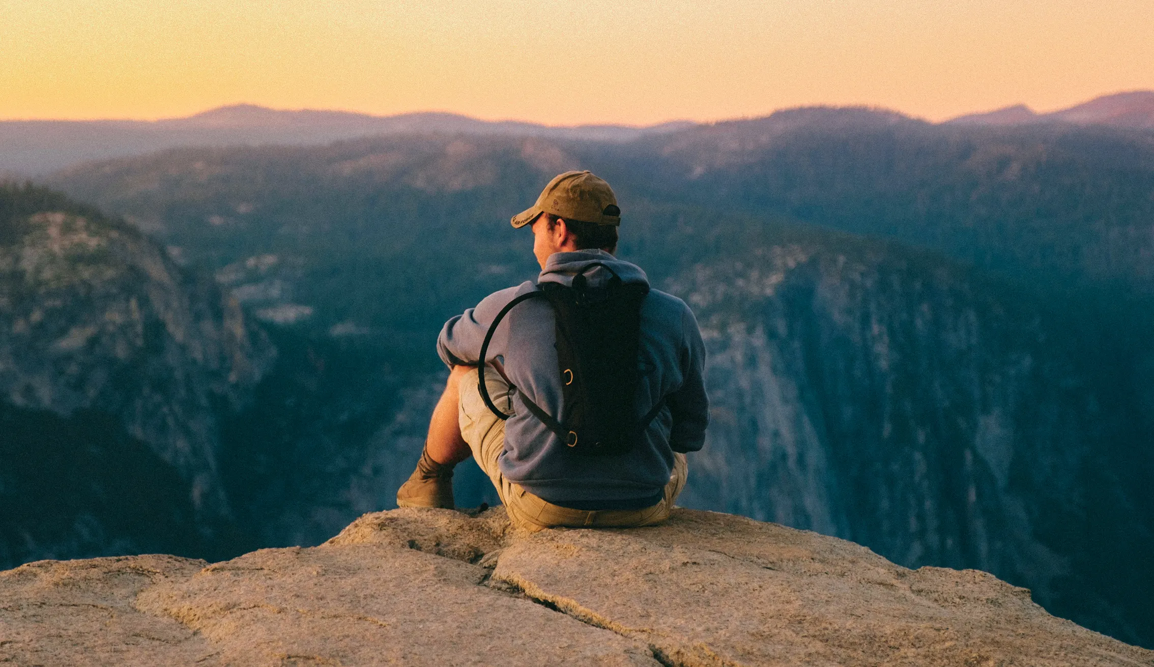 Man wearing a cap and backpack sitting on a rocky cliff overlooking a mountainous landscape at sunset.