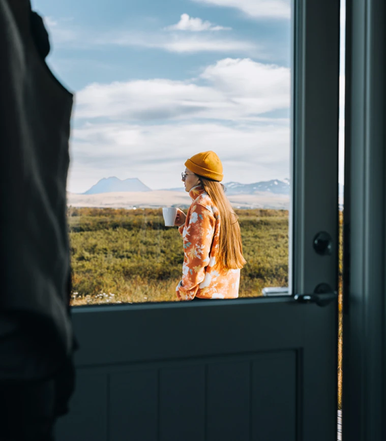 Person in an orange floral jacket and yellow beanie holding a white cup, standing outside with mountains and cloudy sky in the background, viewed through a partially open door.