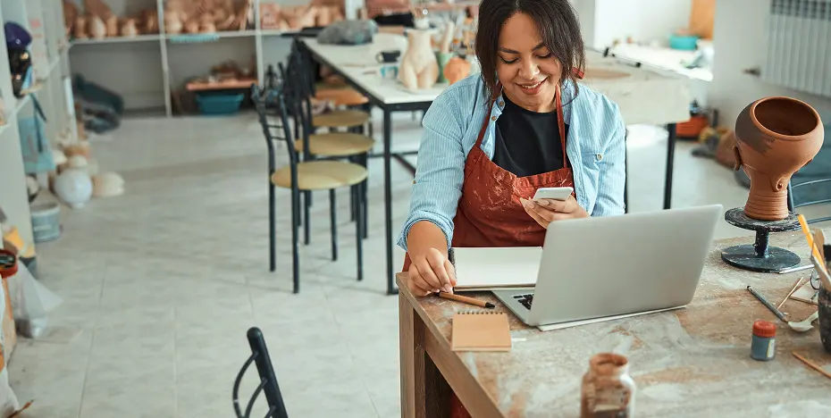 Woman in an apron using a smartphone and laptop at a cluttered pottery studio table.
