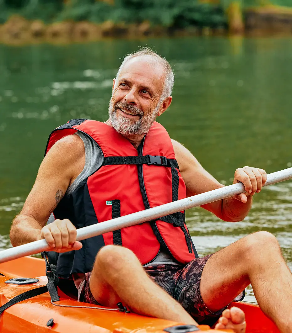 Smiling middle-aged man wearing a red life jacket paddling an orange kayak on calm water.