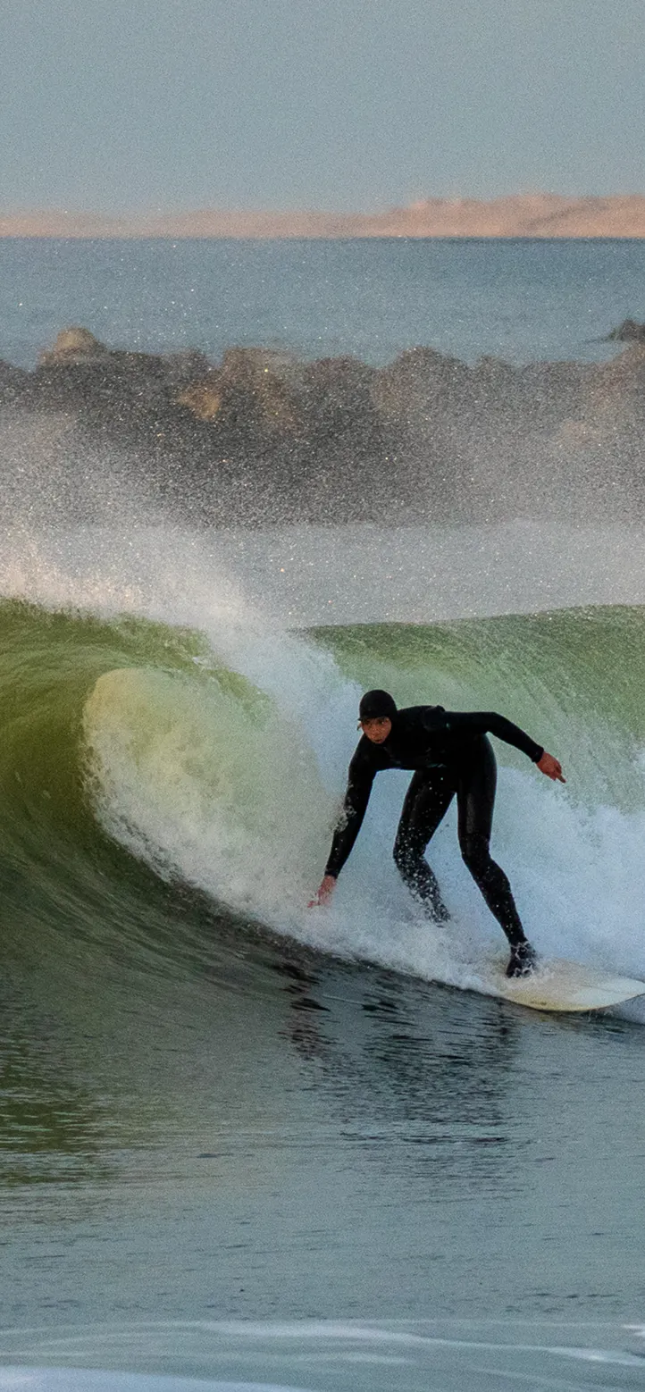 Surfer dressed in a black wetsuit riding a green wave near a rocky breakwater.