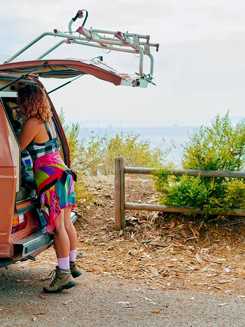 Woman with curly hair and a colorful skirt leaning into the back of an open van near a wooden fence and bushes by a coastal area.