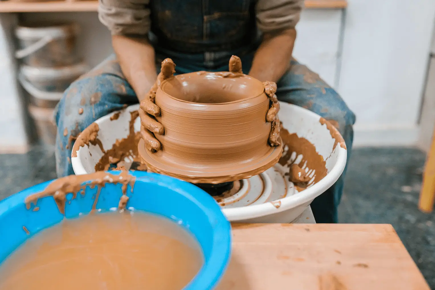 Hands shaping a clay pot on a spinning pottery wheel with a blue basin of water nearby.