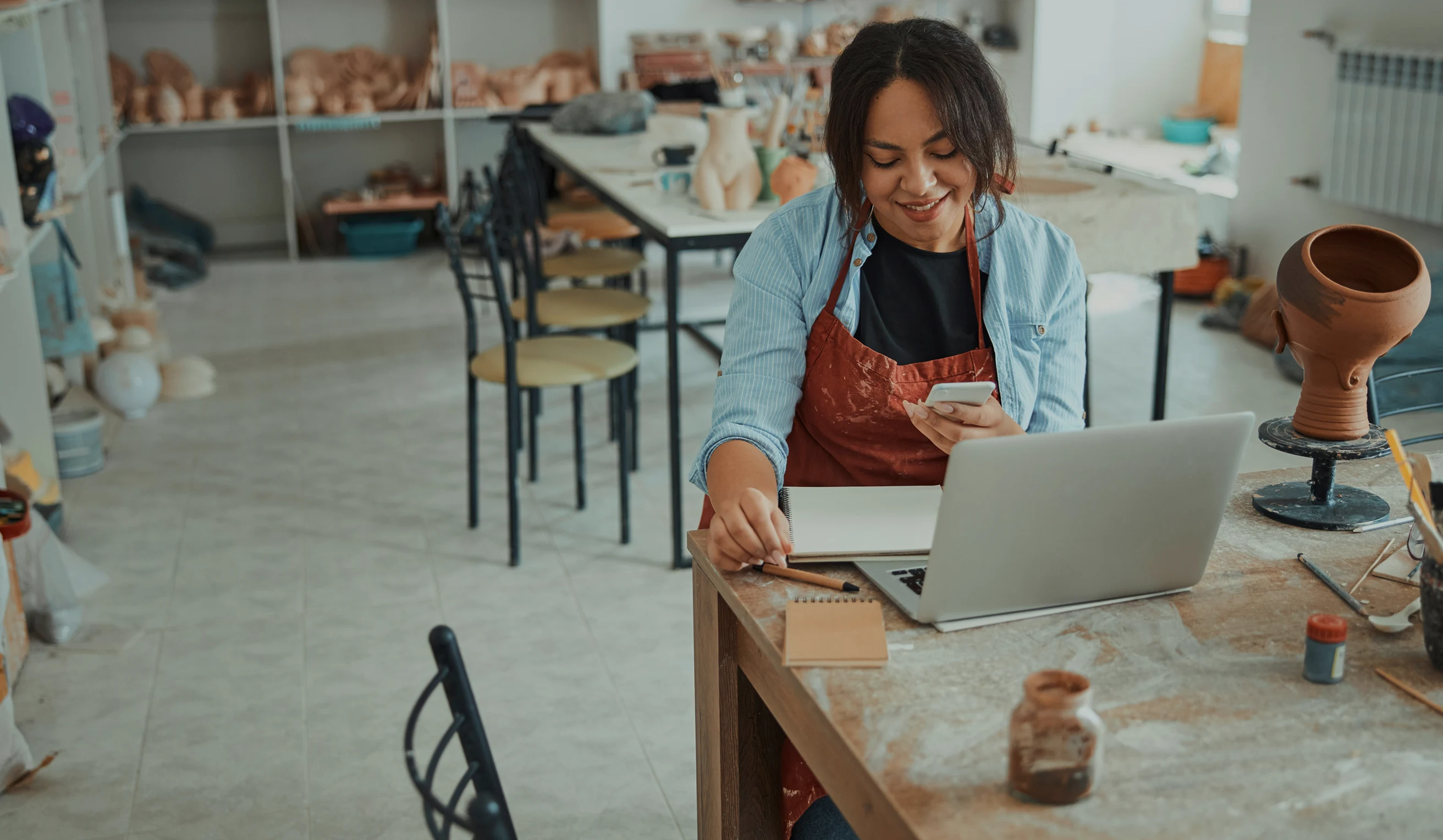 Smiling woman in a red apron sitting at a table using a smartphone and laptop in a pottery studio.