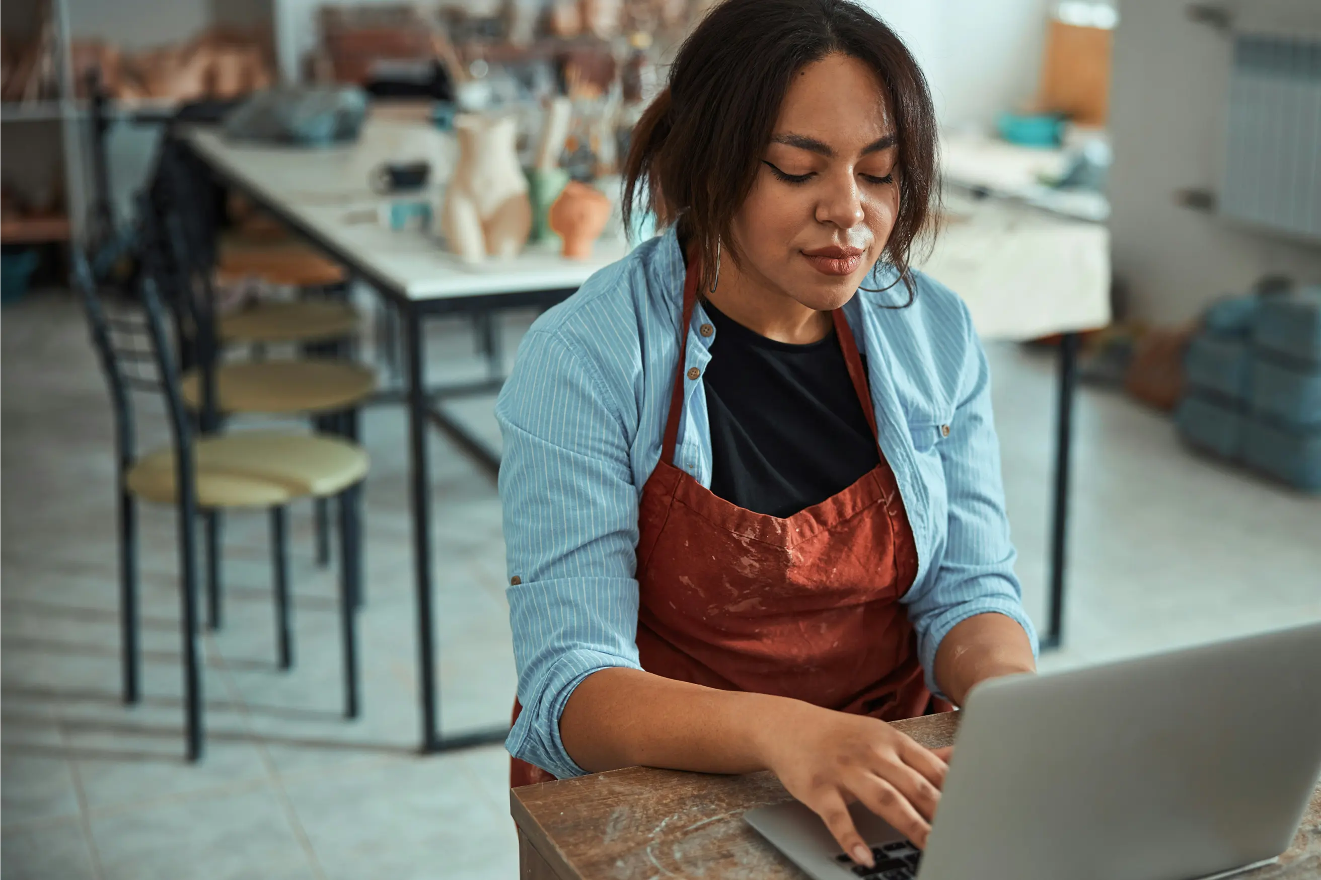 Woman wearing a red apron and blue shirt working on a laptop in a workshop with mannequin torsos in the background.