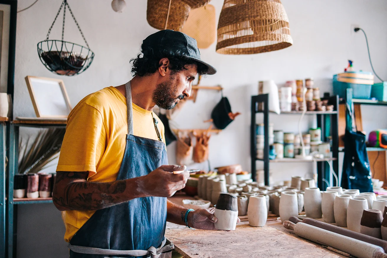 Man in a yellow shirt and apron painting a ceramic cup in a pottery workshop filled with pottery pieces and shelves.