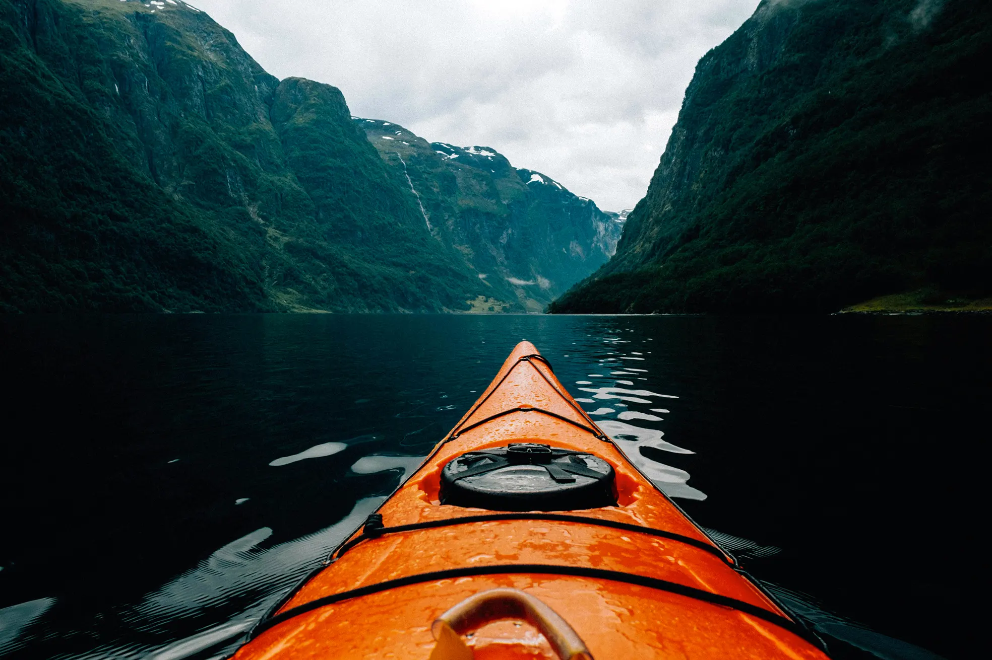 View from an orange kayak on calm water surrounded by green mountains under a cloudy sky.