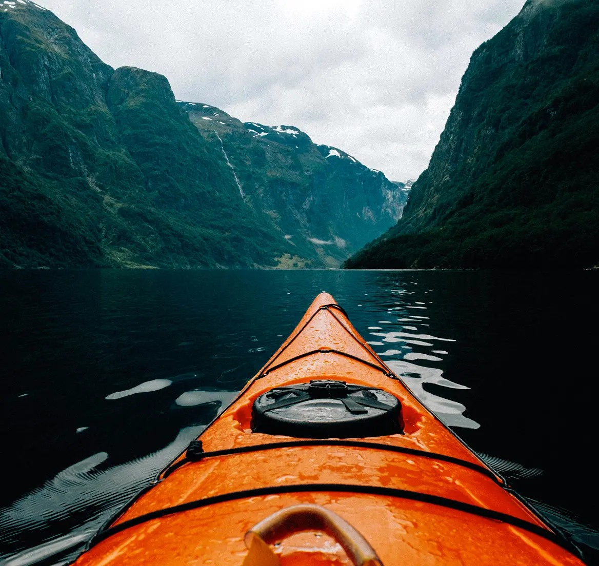 View from an orange kayak on calm water surrounded by tall, green mountains under a cloudy sky.