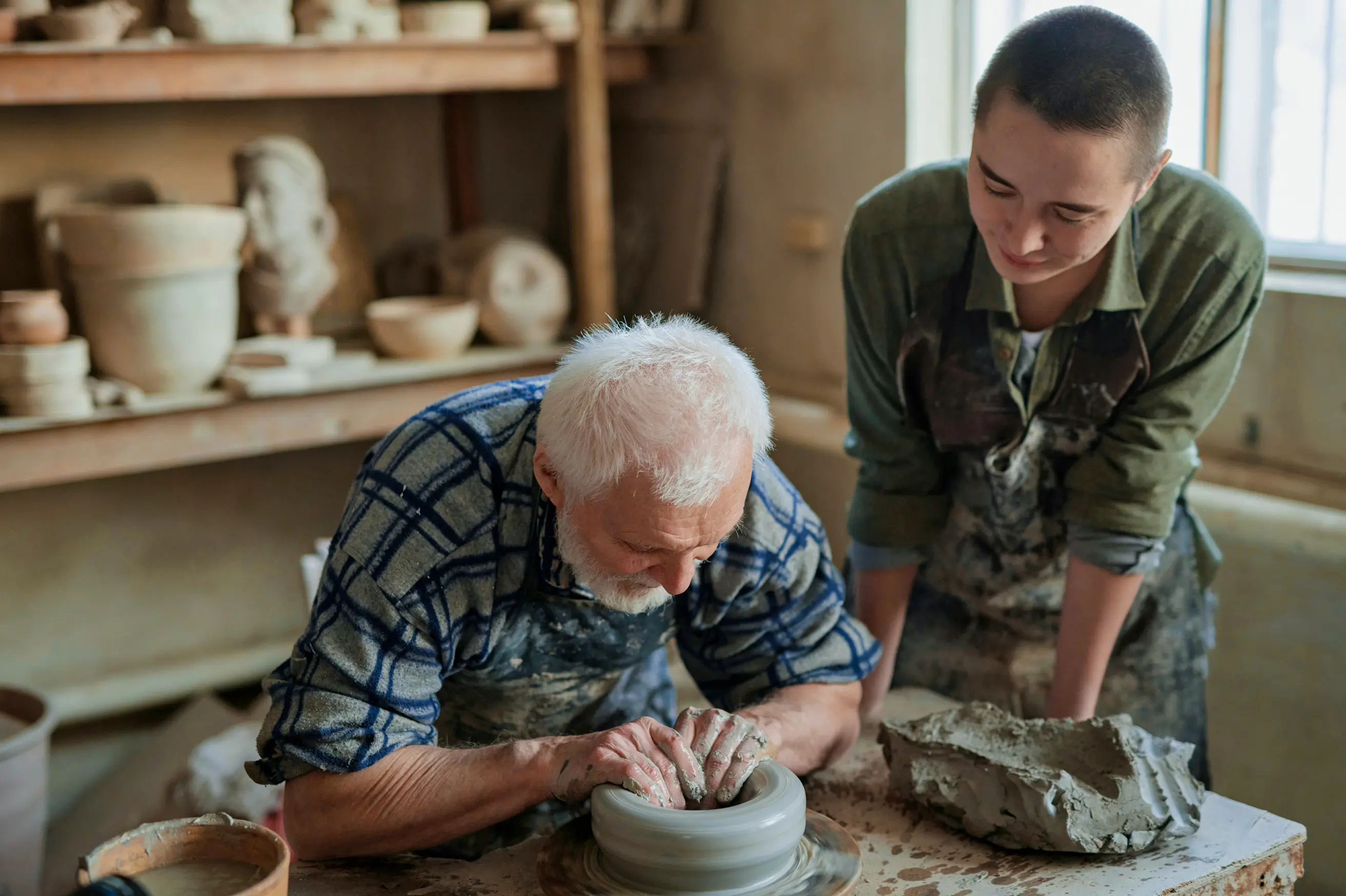 An older man shaping clay on a pottery wheel while a younger person watches and learns in a pottery studio.