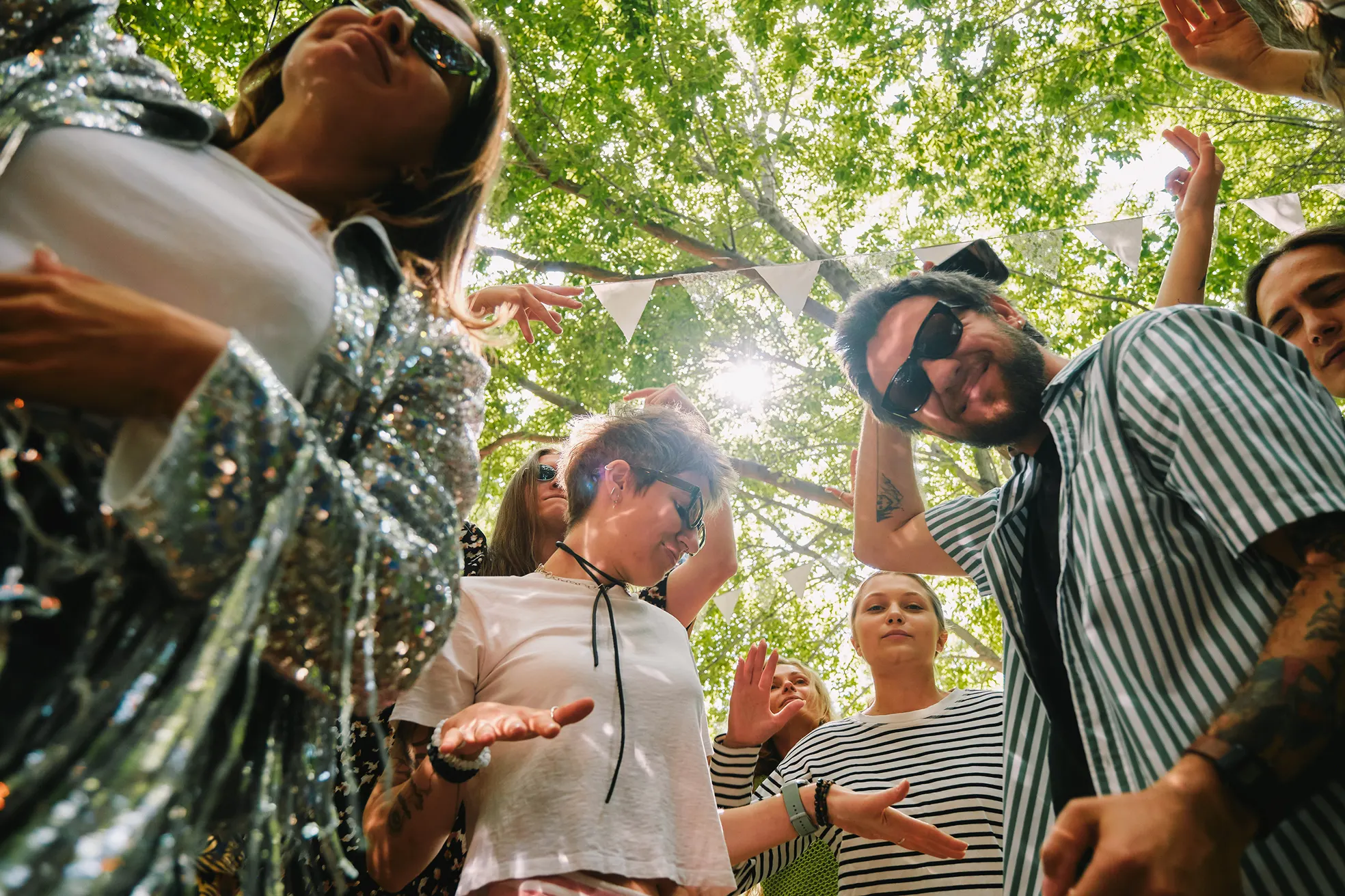 Group of young people wearing sunglasses and casual clothes dancing outdoors under green trees and sunlight.