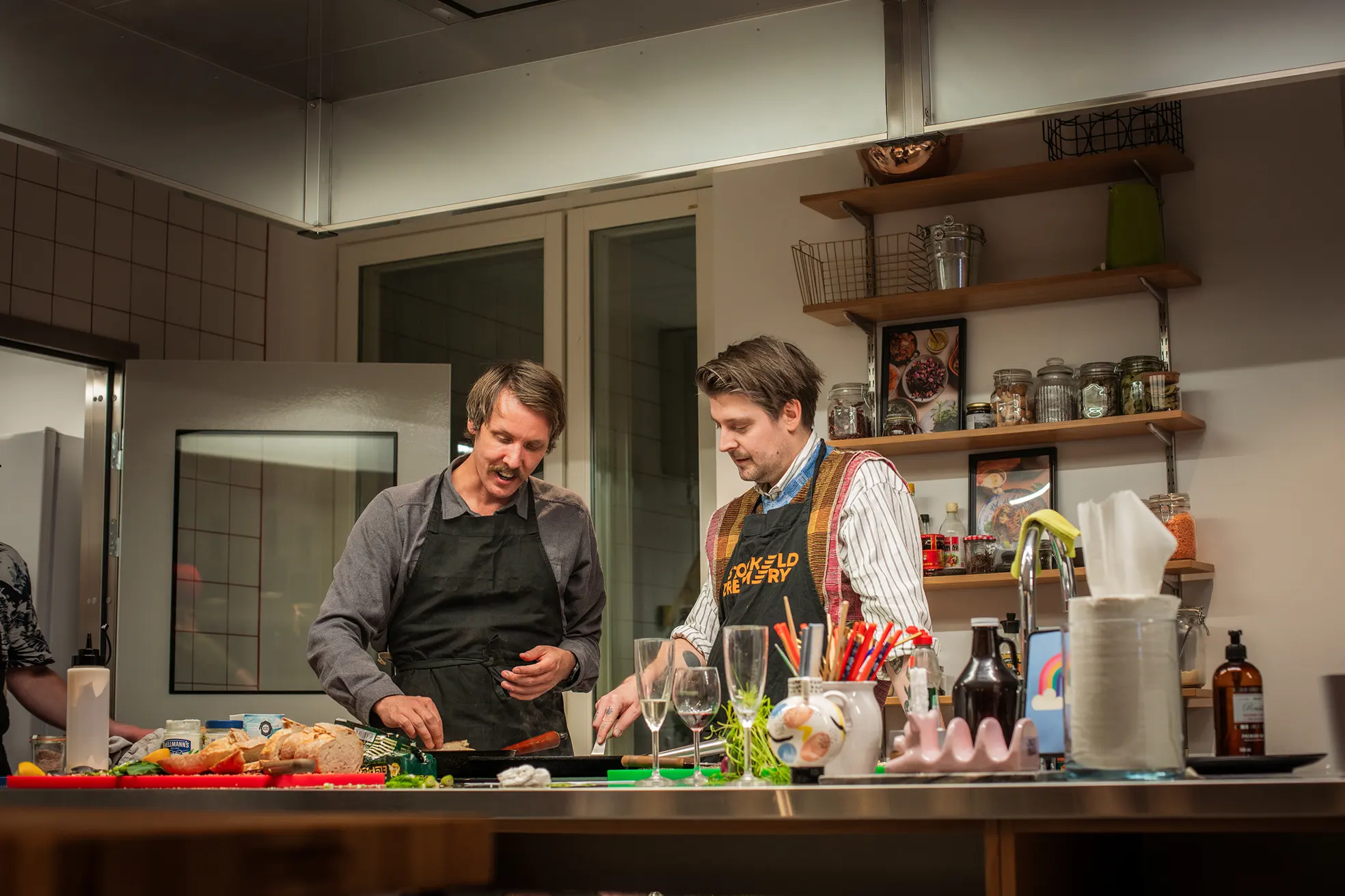 Two men wearing aprons cooking together in a kitchen with shelves of jars and spices behind them.