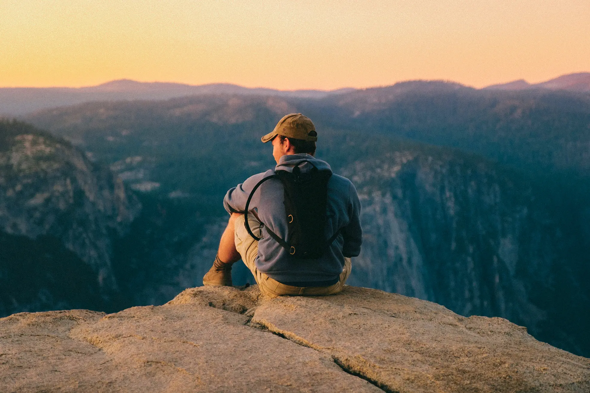 Man wearing a hat and backpack sitting on a rocky cliff overlooking a mountain landscape at sunset.
