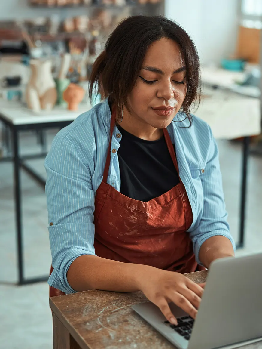 Woman wearing a blue shirt and red apron working on a laptop in an arts and crafts studio.