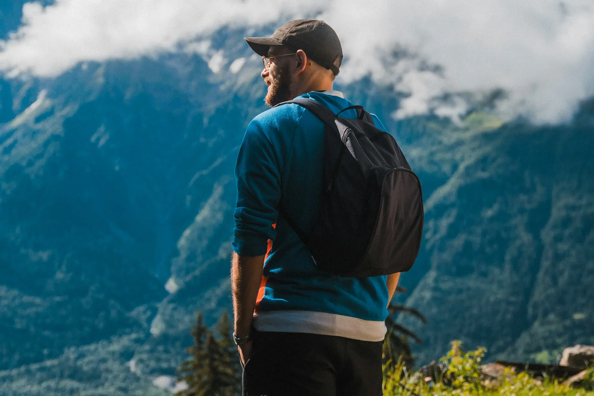 Man wearing a cap and backpack looking at mountainous landscape under a cloudy sky.