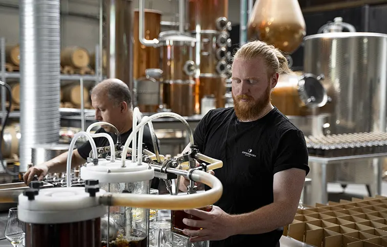 Two men working in a distillery with copper equipment, filling bottles with liquid.