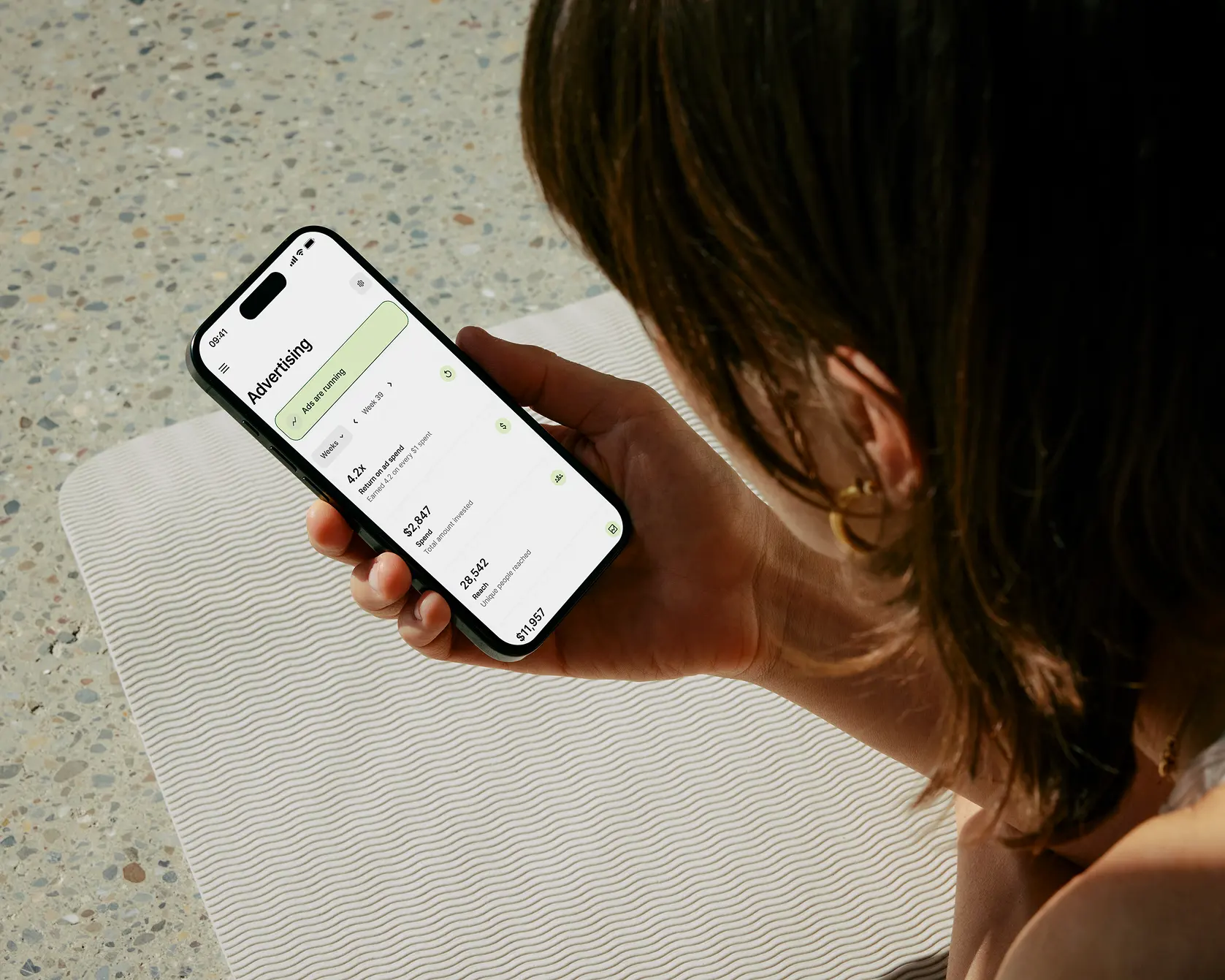 Person holding a smartphone displaying an advertising app screen with metrics like return on ad spend, spend amount, and unique people reached.