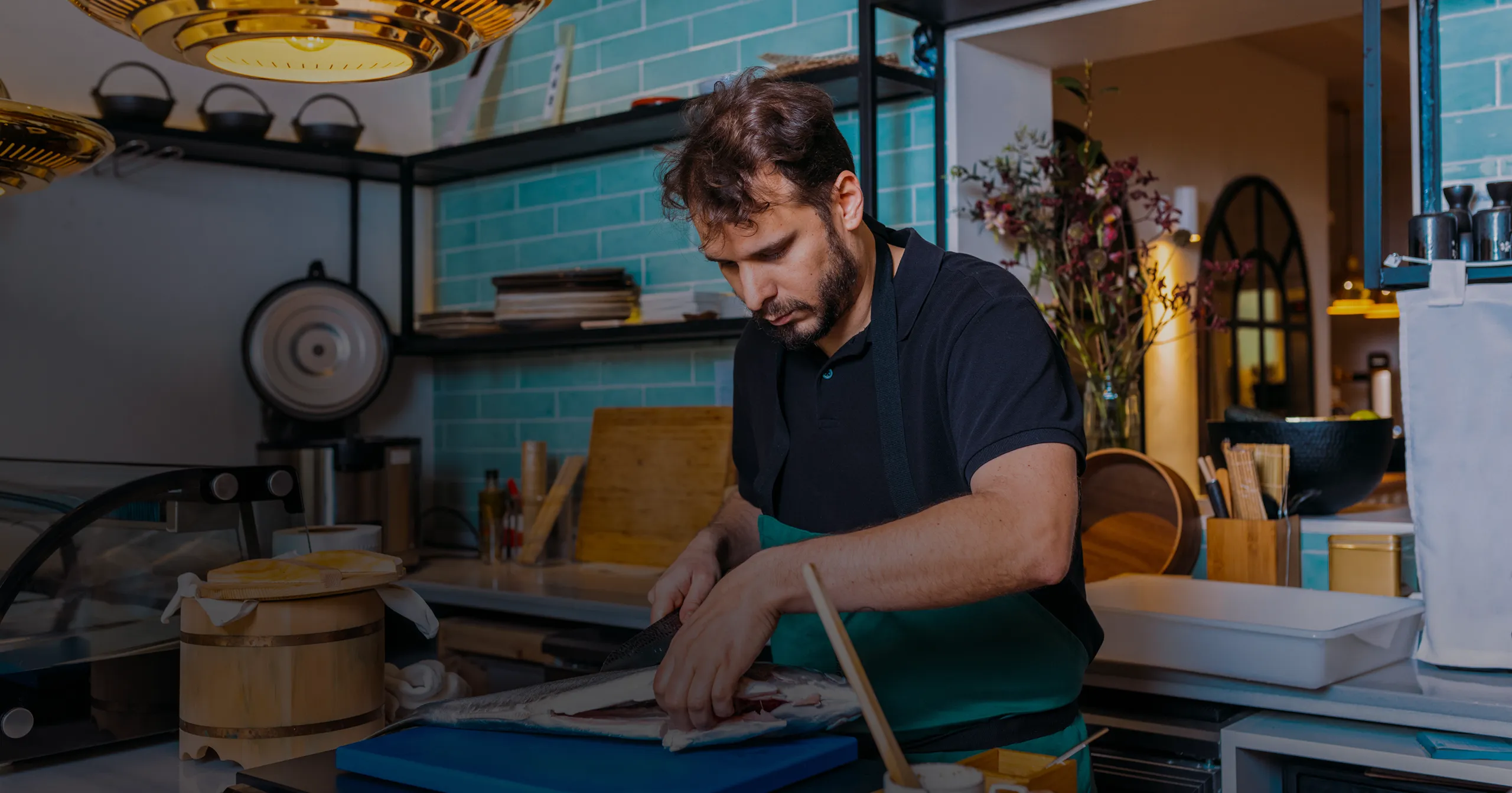 Man in black shirt and apron filleting a large fish on a blue cutting board in a kitchen with teal tiled walls.